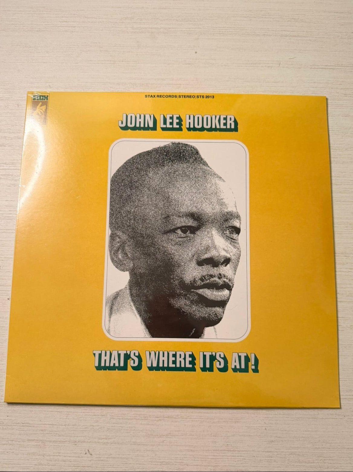 A vinyl record album cover for John Lee Hooker's 'That's Where It's At!' resting on a light-colored wooden surface. The cover features a bright yellow background with a central black and white portrait of the artist and bold black text outlined in teal.